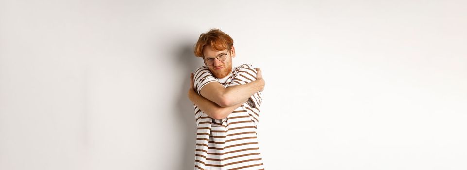Reluctant young man with messy red hair unwilling do something, hugging himself and grimacing, standing over white background.