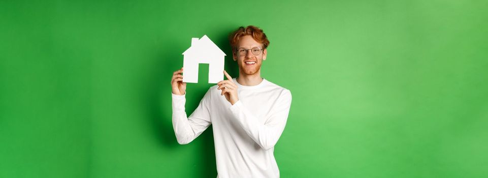 Real estate. Handsome young man with red hair, wearing glasses, showing paper house cutout and smiling, standing against green background.