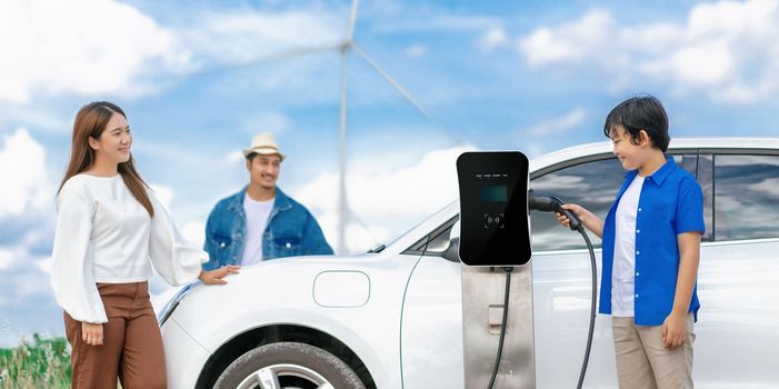 Concept of progressive happy family enjoying their time at wind farm with electric vehicle. Electric vehicle driven by clean renewable energy from wind turbine generator for charging station.