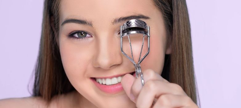 Closeup happy young charming brunette holding eyelash curler in her hand as makeup accessory. Beautiful girl applying eyelash curler in studio with isolated background for copyspace.