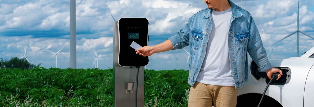 Progressive man with his electric car, EV car recharging energy from charging station on green field with wind turbine as concept of future sustainable energy. Electric vehicle with energy generator.