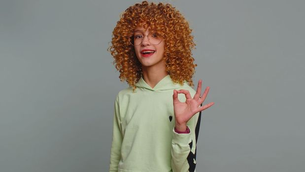 Ok. Young teenager school girl with curly hair looking approvingly at camera showing okey gesture, like sign positive, approve something good. Female fit child kid isolated on studio gray background