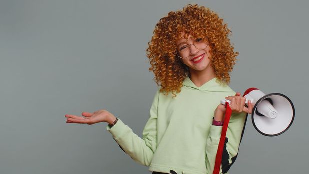 Young teenager girl in glasses scream shout in megaphone loudspeaker announces discounts sale, announcing advertisement pointing empty place area. Child kid isolated with curly hair on gray background