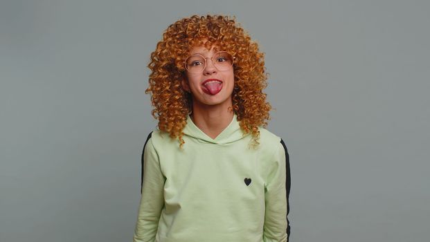 Cheerful bully young teenager girl in glasses showing tongue making faces at camera, fooling around, joking, aping with silly face, teasing you. Curly hair child kid on gray background. Funny children