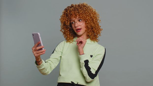 Young child girl kid blogger taking selfie on smartphone holding in hand, communicating video call talking online with subscribers. Teenager children with curly hair isolated on studio gray background