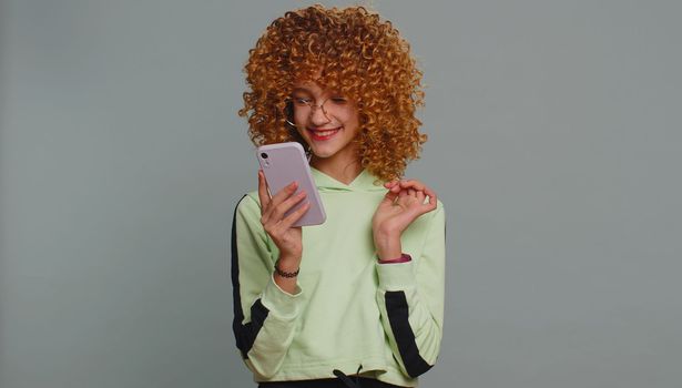 Young child girl kid with curly hair use mobile smartphone typing browsing say Wow yes found out great big win good news celebrate doing winner gesture. Teenager children on studio gray background