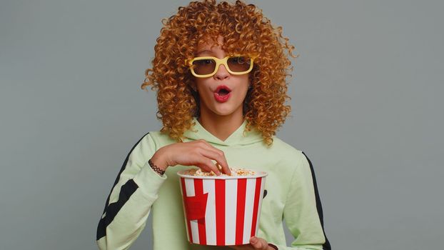 Excited young teenager school girl in 3D glasses eating popcorn, watching interesting tv serial, sport game, film, online social media movie content. Curly haired child kid on studio gray background
