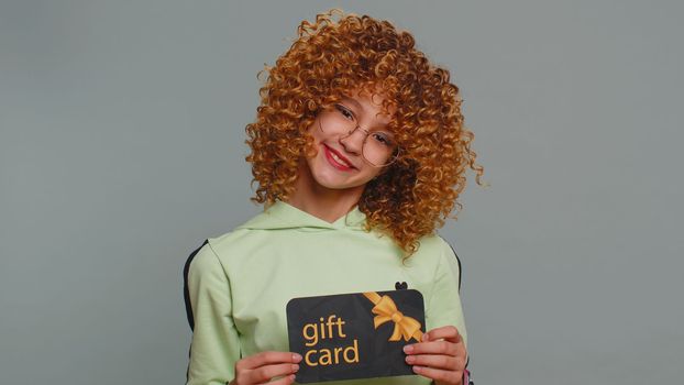 Teenager school girl showing pointing on gift discount certificate voucher coupon for store holidays sale. Child kid with shopping christmas surprise card isolated alone on gray studio background