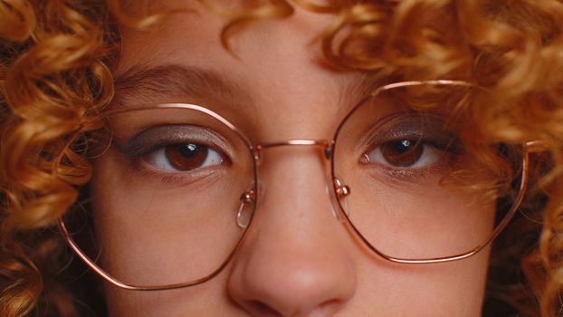 Extreme close-up macro portrait of smiling girl face. Teen beautiful kid's eyes, looking at camera. Brown eyes of redhead female child model. Young children in eyeglasses opening wide her closed eyes
