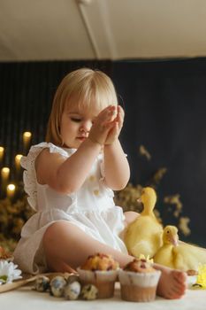 A little girl is sitting on the Easter table and playing with cute fluffy ducklings. The concept of celebrating happy Easter