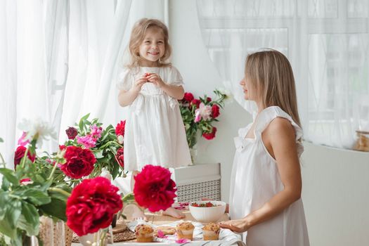 A little blonde girl with her mom on a kitchen countertop decorated with peonies. The concept of the relationship between mother and daughter. Spring atmosphere.