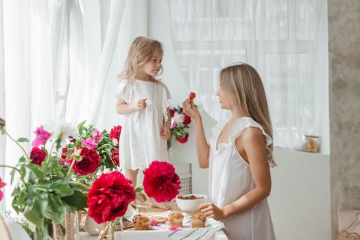 A little blonde girl with her mom on a kitchen countertop decorated with peonies. The concept of the relationship between mother and daughter. Spring atmosphere.