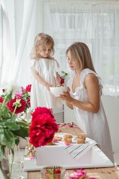 A little blonde girl with her mom on a kitchen countertop decorated with peonies. The concept of the relationship between mother and daughter. Spring atmosphere.