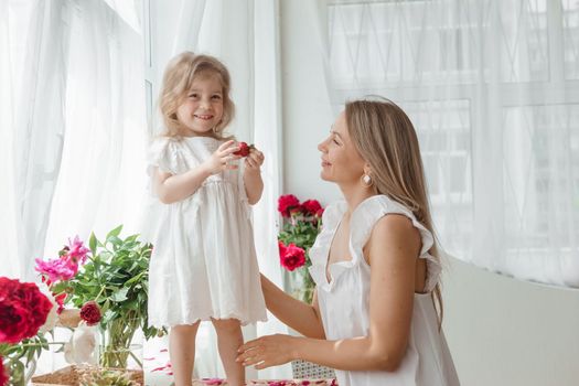 A little blonde girl with her mom on a kitchen countertop decorated with peonies. The concept of the relationship between mother and daughter. Spring atmosphere.