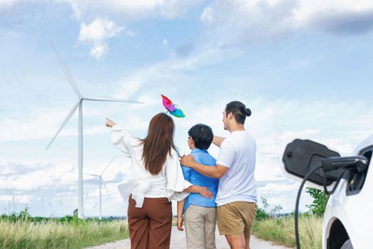 Concept of progressive happy family holding windmill toy and relax at wind farm with electric vehicle. Electric vehicle driven by clean renewable energy from wind turbine generator to charger station.