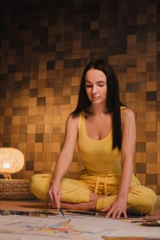 A girl in yellow clothes sitting in the Lotus position draws on paper with evening lighting at home.