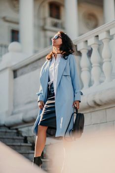 Portrait of a brunette business lady with glasses. Woman walks around the city, lifestyle. She wears a blue cloak, a black leather skirt, and black heeled boots