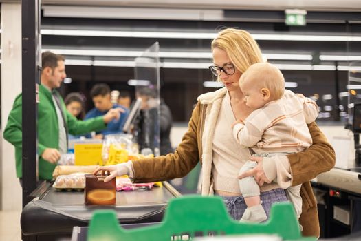 Mother shopping with her infant baby boy, holding the child while stacking products at the cash register in supermarket grocery store