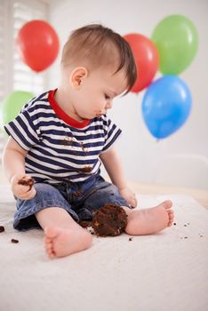 Hes sampling his birthday cake. a baby boy sitting at home with balloons in the background