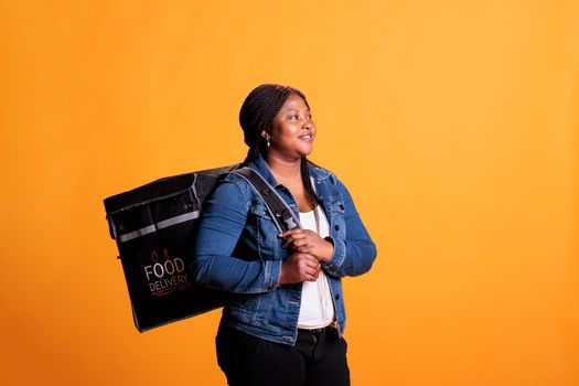 Smiling restaurant courier standing in studio over yellow background carrying takeaway thermal backpack ready to deliver meal order to customer. Food transportation service and takeout concept