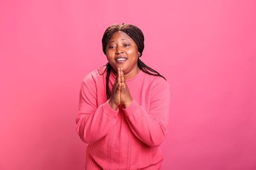 Happy female person doing prayer hands sign in studio, showing religious spiritual gesture over pink background. Young model with confidence praying to jesus for peace, christianity.