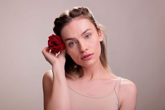 Attractive caucasian woman putting red rose in blonde hair face portrait. Beautiful young lady holding bright flower bud behind ear and looking at camera with romantic glance
