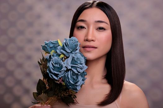 Young woman holding blue roses bouquet near face portrait. Natural beautiful asian model with nude make up posing with elegant flowers branch in arms and looking at camera