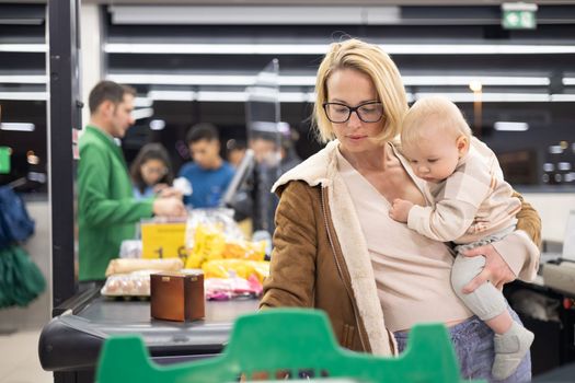 Mother shopping with her infant baby boy, holding the child while stacking products at the cash register in supermarket grocery store