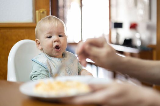 Mother spoon feeding her infant baby boy child sitting in high chair at the dining table in kitchen at home.