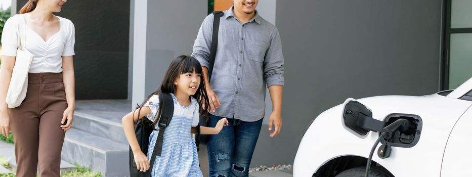 Progressive young parents and daughter with electric vehicle and home charging station. Green and clean energy from electric vehicles for healthy environment. Eco power from renewable source at home.