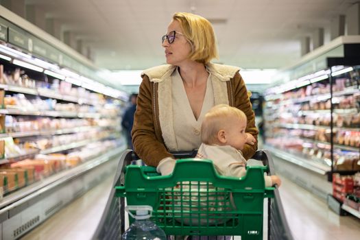 Mother pushing shopping cart with her infant baby boy child down department aisle in supermarket grocery store. Shopping with kids concept