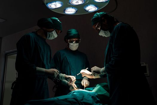 Surgical team performing surgery to patient in sterile operating room. In a surgery room lit by a lamp, a professional and confident surgical team provides medical care to an unconscious patient.