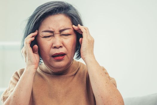 An agonizing senior woman with a headache, compress her temple with both hands at her contented living room. Senior health, nursing home, caretaker service.