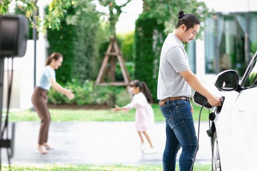 Focus image of progressive man charging electric car from home charging station with blur mother and daughter playing together in the background.