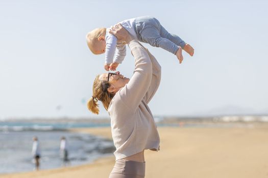 Mother enjoying summer vacations holding, playing and lifting his infant baby boy son high in the air on sandy beach on Lanzarote island, Spain. Family travel and vacations concept