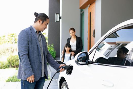 Progressive young parents and daughter with electric vehicle and home charging station. Green and clean energy from electric vehicles for healthy environment. Eco power from renewable source at home.