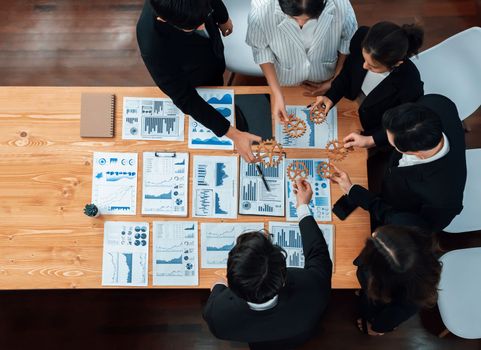 Top view hand holding gear by businesspeople wearing suit for harmony synergy in office workplace concept. Group of people hand making chain of gears into collective form with dashboard report papers.