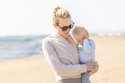 Mother holding and carrying his infant baby boy son on sandy beach enjoying summer vacationson on Lanzarote island, Spain. Family travel and vacations concept