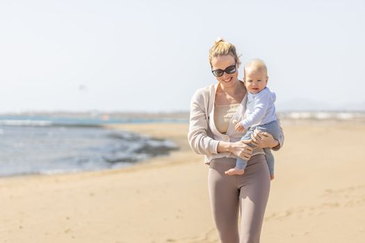 Mother holding and carrying his infant baby boy son on sandy beach enjoying summer vacationson on Lanzarote island, Spain. Family travel and vacations concept