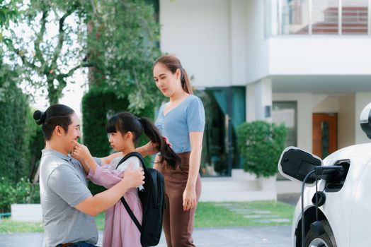 Progressive young parents and daughter with electric vehicle and home charging station. Green and clean energy from electric vehicles for healthy environment. Eco power from renewable source at home.