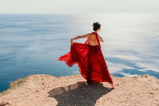 Side view a Young beautiful sensual woman in a red long dress posing on a rock high above the sea during sunrise. Girl on the nature on blue sky background. Fashion photo.