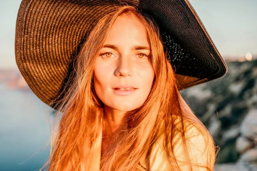 Portrait of happy young woman wearing summer black hat with large brim at beach on sunset. Closeup face of attractive girl with black straw hat. Happy young woman smiling and looking at camera at sea