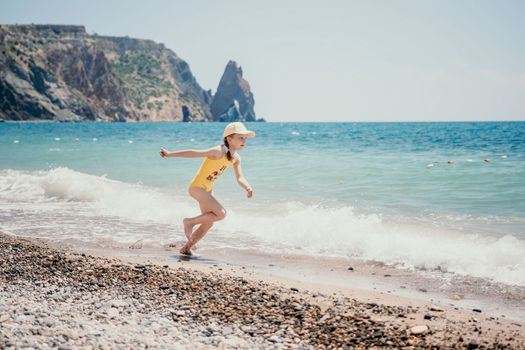 Cute little girl running along the seashore against a clear blue sea and rejoices in the rays of the summer sun. Beautiful girl in yellow swimsuit running and having fun on tropical beach