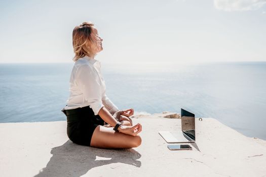 Happy girl doing yoga with laptop working at the beach. beautiful and calm business woman sitting with a laptop in a summer cafe in the lotus position meditating and relaxing. freelance girl remote work beach paradise