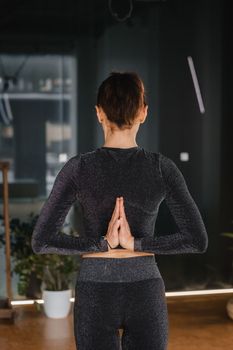 A girl in a black sports uniform does yoga standing on a mat in the gym.