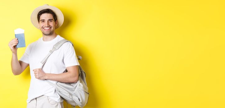 Tourism and vacation. Smiling young guy going on trip, holding backpack and showing passport with tickets, standing over yellow background.