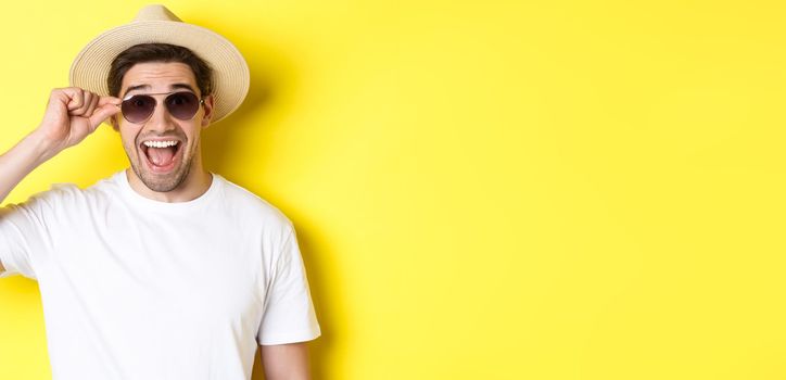 Concept of tourism and holidays. Close-up of happy man in summer hat and sunglasses enjoying vacation, standing over yellow background.