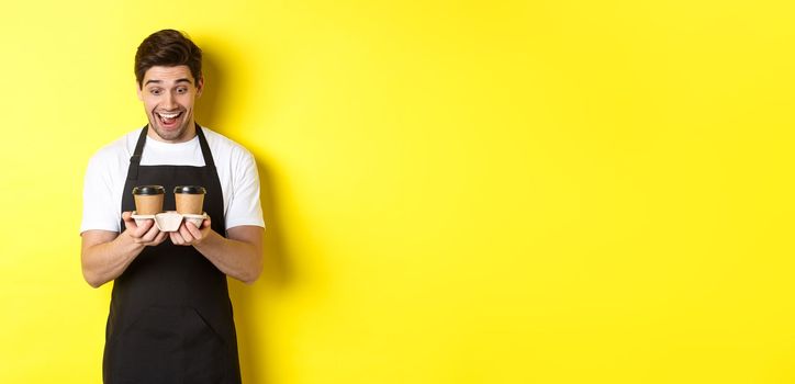 Waiter looking excited at two cups of takeaway coffee, wearing black apron, standing over yellow background.