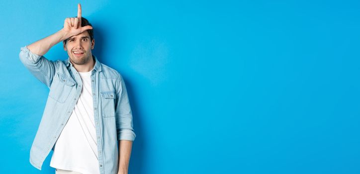 Rude guy mocking person for losing, showing loser sign on forehead and looking with dismay, standing against blue background.