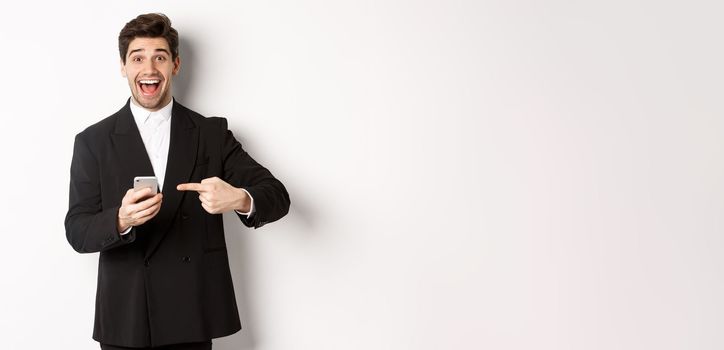 Image of cheerful businessman looking amazed, pointing at mobile phone, standing in suit over white background.